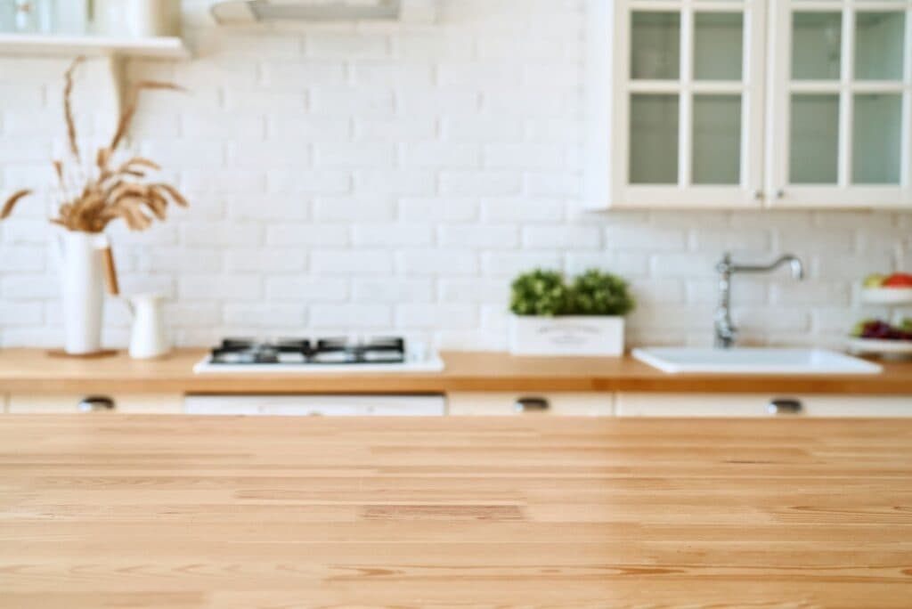 Blurred modern kitchen interior with wooden countertop, white cabinets, brick backsplash, sink, and potted plants in the background