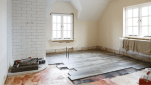 Bright, partially renovated room with white walls and unfinished wood flooring. Two large windows provide natural light. Tools are scattered on the floor.