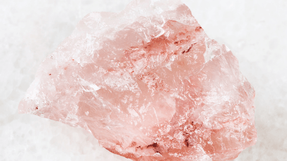 A large raw rose quartz crystal with a soft pink hue and rough texture, set against a light background. It conveys a sense of natural beauty and tranquility.