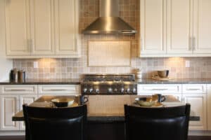 Elegant kitchen with beige cabinets and backsplash, stainless steel range and hood. A dark island has two bowls, mugs, and chairs, creating a cozy feel.