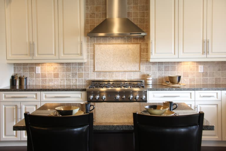 Elegant kitchen with beige cabinets and backsplash, stainless steel range and hood. A dark island has two bowls, mugs, and chairs, creating a cozy feel.