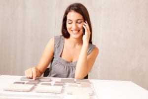 Smiling woman on phone at table, examining tile samples. She wears a gray sleeveless top, conveying a sense of focus and decision-making.