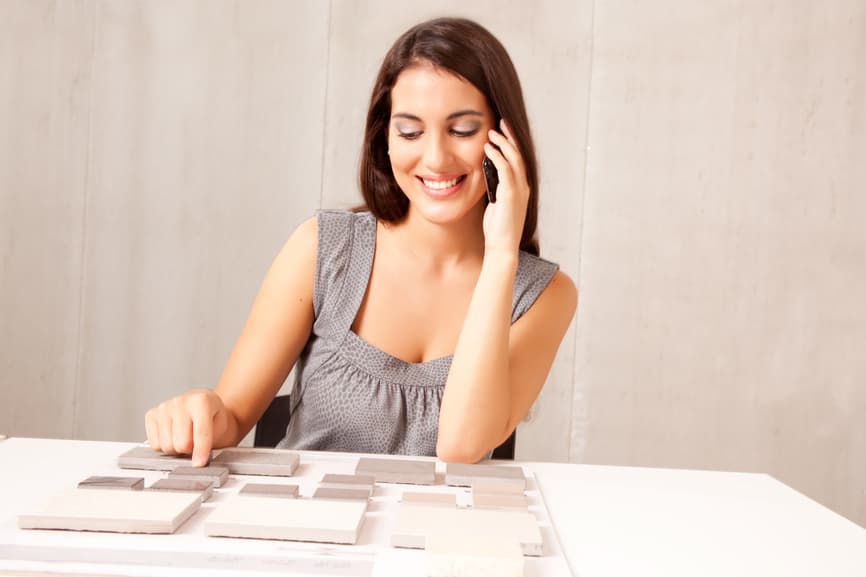 Smiling woman on phone at table, examining tile samples. She wears a gray sleeveless top, conveying a sense of focus and decision-making.
