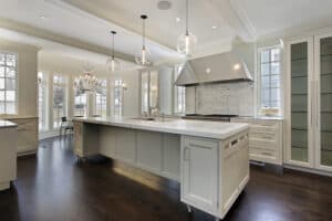 Spacious modern kitchen with an island, marble countertops, pendant lights, wood floors, and a chandelier. Bright daylight fills the room through tall windows.