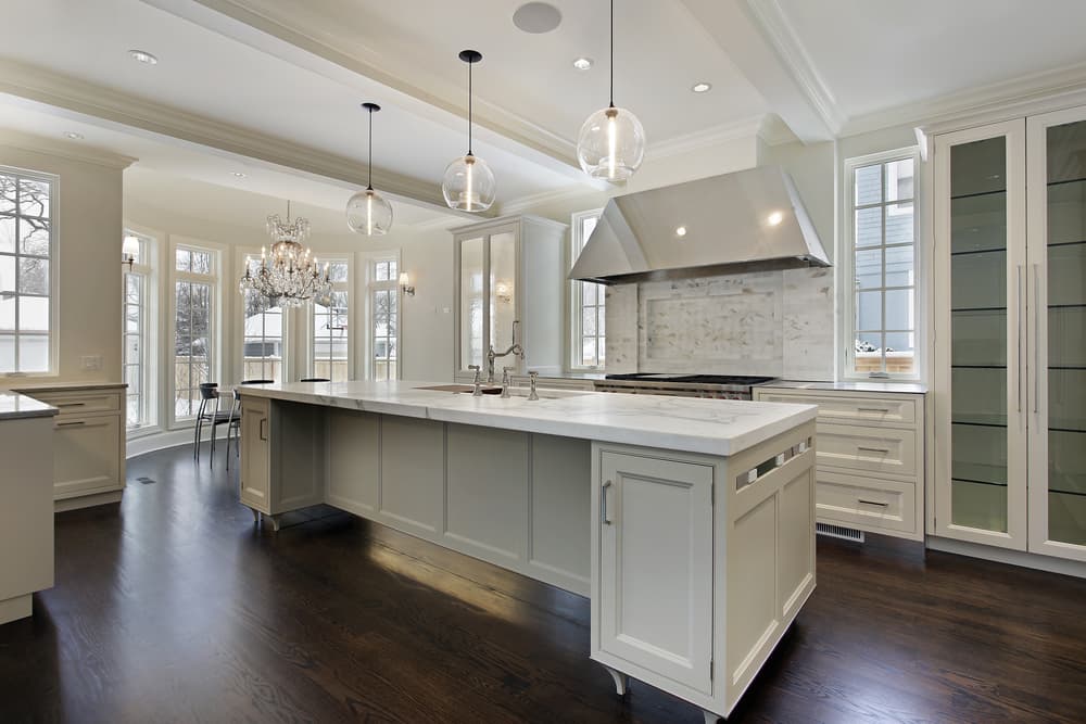 Spacious modern kitchen with an island, marble countertops, pendant lights, wood floors, and a chandelier. Bright daylight fills the room through tall windows.