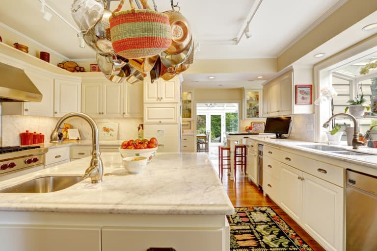 Bright, spacious kitchen with marble countertops, hanging pots, and sleek white cabinets. A bowl of apples adds a pop of color. Large windows illuminate the room.