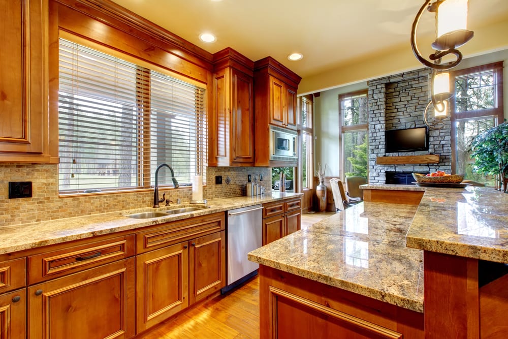 Warm, spacious kitchen with wooden cabinets and granite countertops, under recessed lighting. A large window overlooks a lush, green backyard. Cozy stone fireplace and TV visible in adjacent living area.