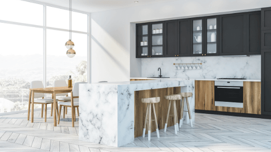 Modern kitchen with white marble island, wooden stools, and dark cabinets. Dining area with wooden table and chairs by large windows, creating a bright and airy feel.