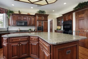 Spacious kitchen with rich wooden cabinets, large granite island, and modern appliances. Inviting and elegant with warm tones and decorative greenery.