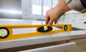 A hand using a yellow spirit level on a speckled kitchen countertop checks alignment. The scene is well-lit, with white cabinets and modern decor.