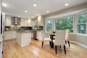 Bright, modern kitchen with white cabinets, granite countertops, and a central island. Hardwood floors extend to a dining area with a round table, surrounded by four beige chairs near large bay windows, offering a view of lush greenery.