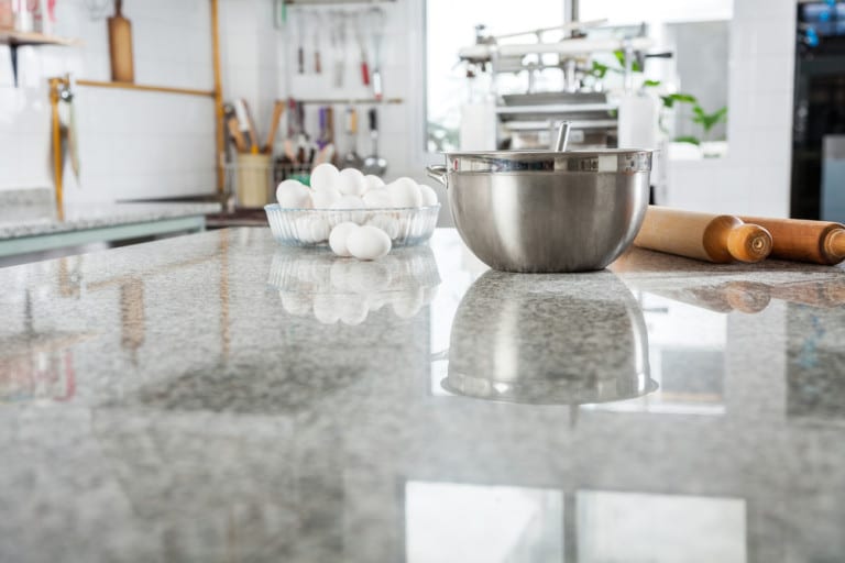 A modern kitchen with a reflective granite countertop, featuring a metal bowl, a rolling pin, and a glass bowl filled with eggs. The ambiance is clean and organized.