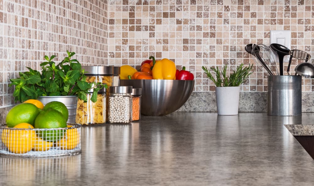 A modern kitchen counter with fresh lemons, limes, herbs, and colorful bell peppers in bowls. Jars of pasta and beans, utensils in a metal holder. Warm, cozy tone.