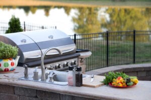 Outdoor kitchen with a silver grill, sink, and condiments. Vibrant plate of vegetables and herbs on a counter, set against a serene lakeside backdrop.