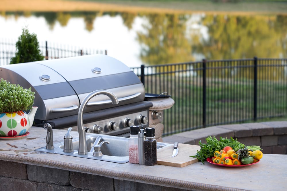 Outdoor kitchen with a silver grill, sink, and condiments. Vibrant plate of vegetables and herbs on a counter, set against a serene lakeside backdrop.