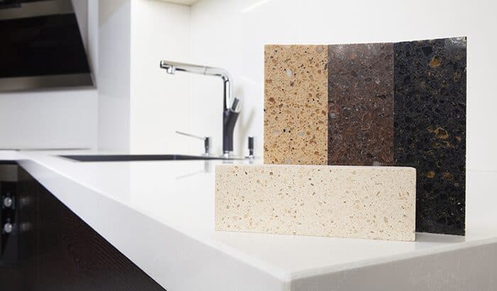 Modern kitchen countertop displaying samples of three stone tiles in beige, brown, and black hues. Sleek faucet in the background.