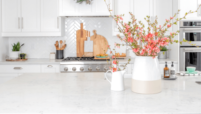 A bright, modern kitchen with white cabinets and a marble countertop. A vase with pink blossoms adds a fresh touch, while wooden cutting boards and plants enhance warmth.