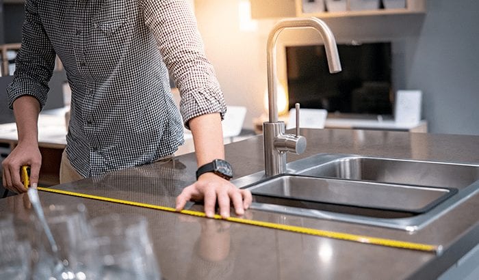 A men measuring a sleek kitchen countertop with a tape measure. The ambient light and modern faucet bring a professional, focused tone.