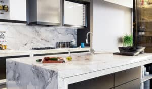 Modern kitchen with a marble island, featuring a cutting board with herbs and oil. Sleek cabinets and a plant add a stylish, minimalist vibe.