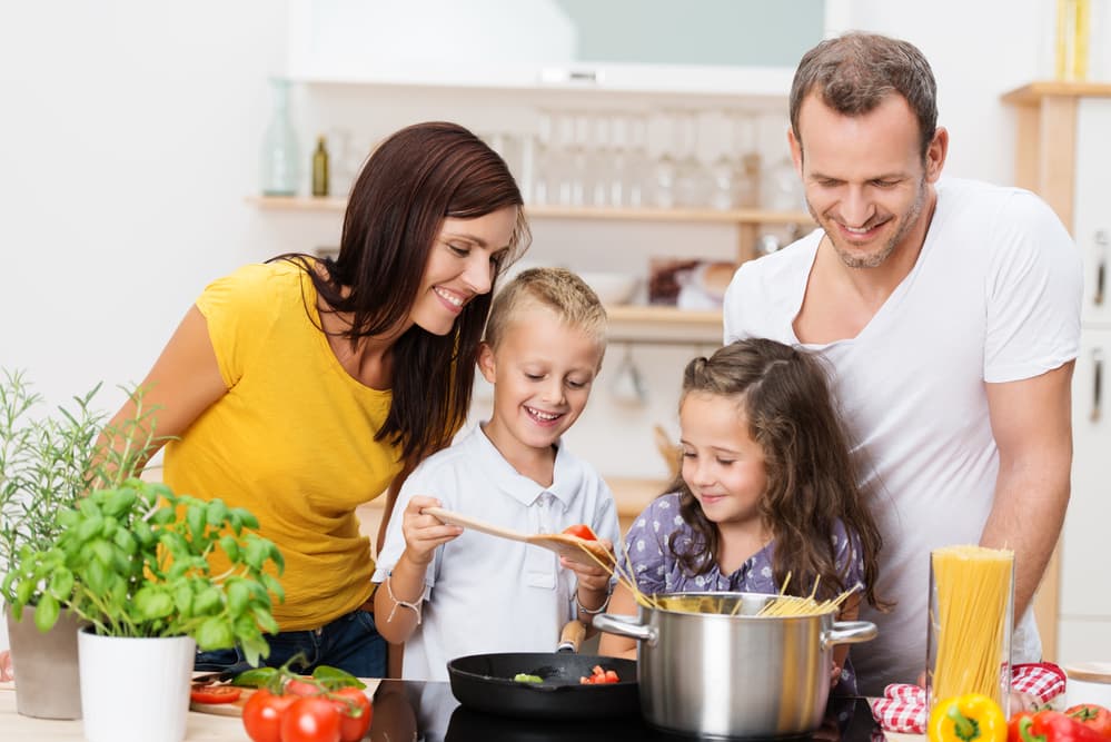 A happy family of four cooks together in a bright kitchen. Two children stand between their smiling parents, holding a ladle over a pot of spaghetti. Fresh vegetables are on the counter.
