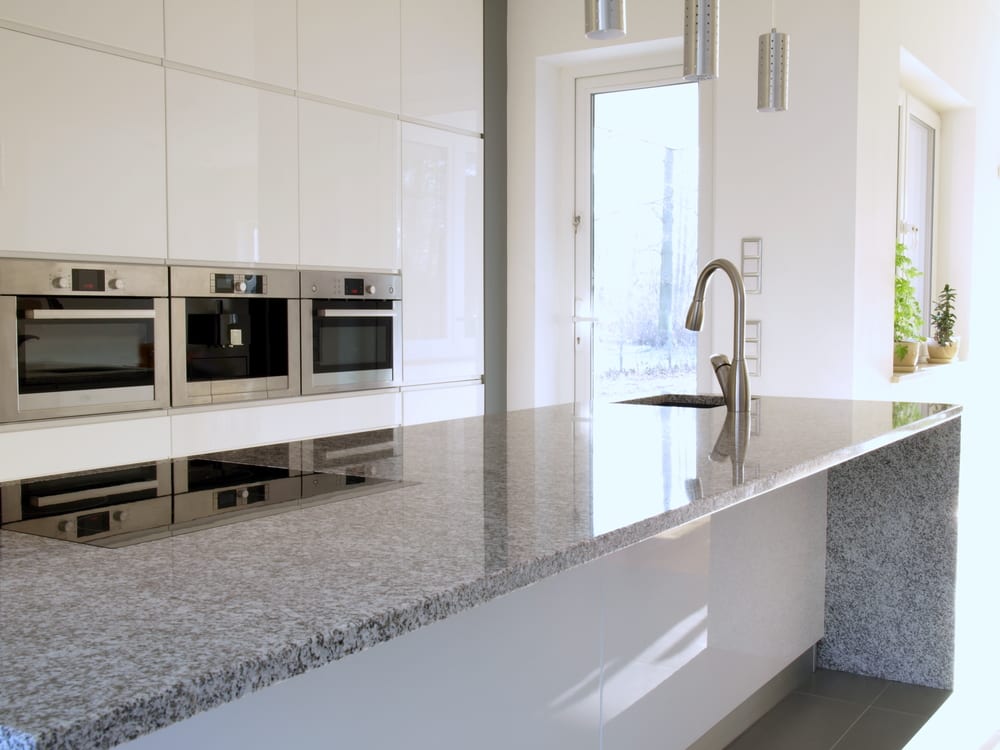 Modern kitchen with sleek white cabinets, stainless steel built-in ovens, and a large granite island with a stainless sink, lit by natural light.