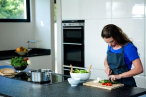 A woman in a blue shirt and striped apron chops vegetables on a wood cutting board in a modern kitchen. There's a salad bowl and cookware on the countertop.