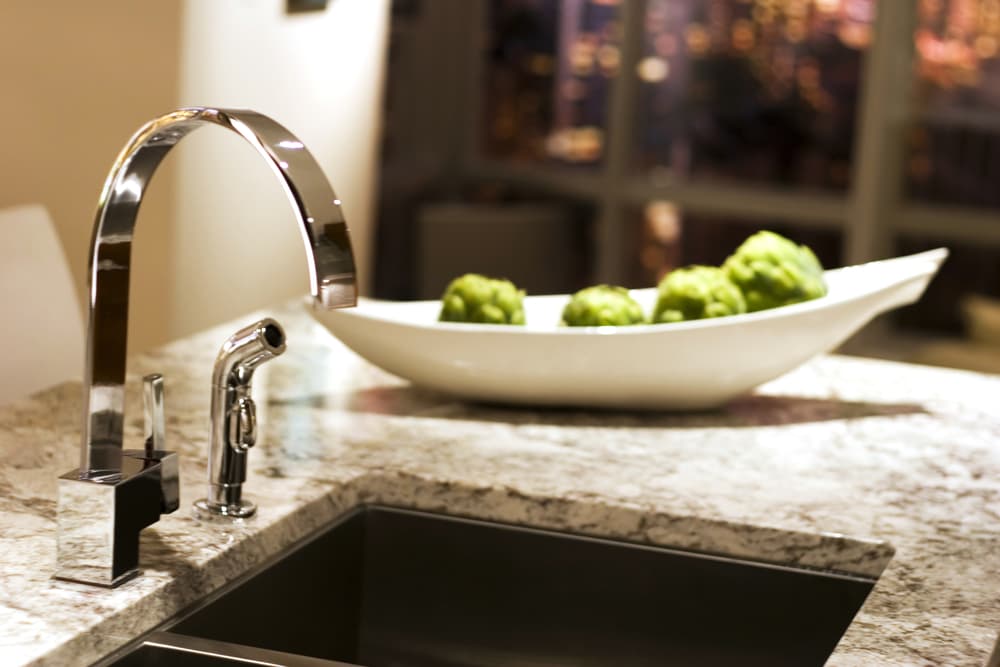 Modern kitchen sink with a sleek chrome faucet on a granite countertop. A white bowl with green artichokes is blurred in the background, creating a contemporary ambiance.