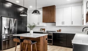 Modern kitchen with sleek black and white cabinetry, stainless steel appliances, and wood accents. An island with pendant lights and stools adds warmth.