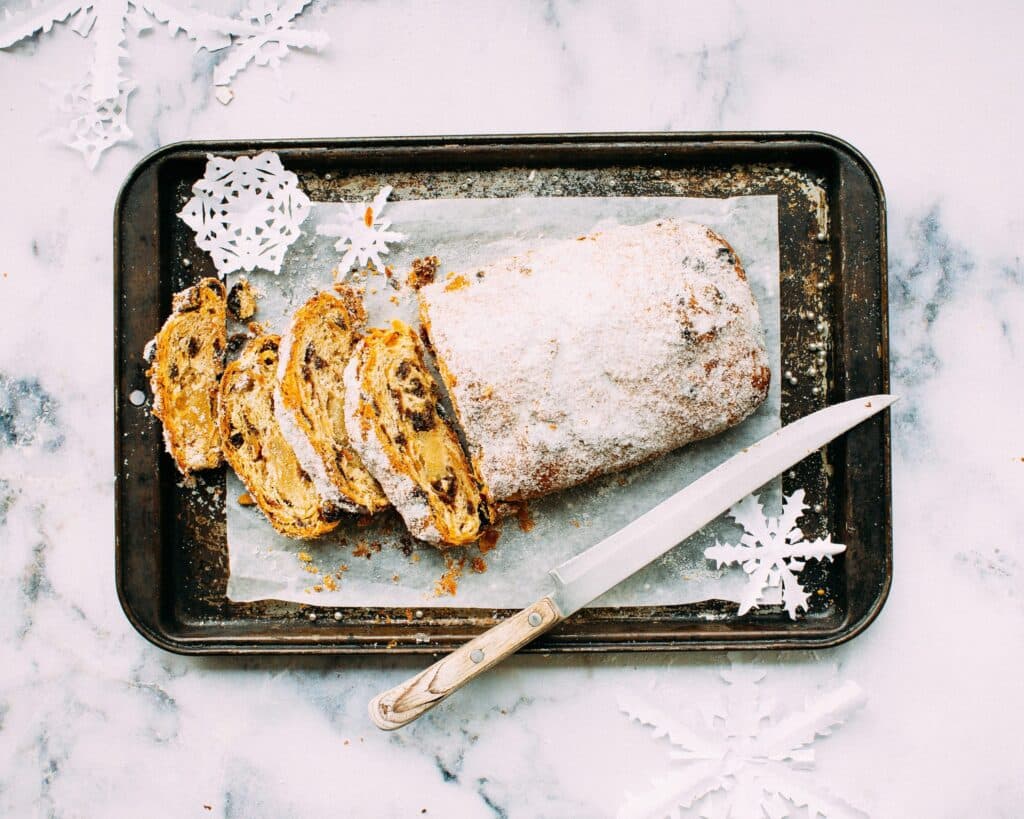 A sliced loaf of sugar-dusted stollen on a baking tray with a knife, surrounded by white paper snowflakes, evoking a festive winter feel.