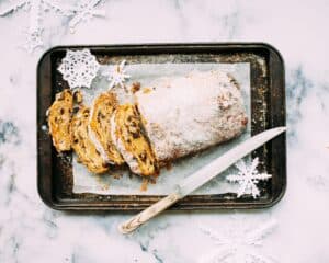 A sliced loaf of sugar-dusted stollen on a baking tray with a knife, surrounded by white paper snowflakes, evoking a festive winter feel.