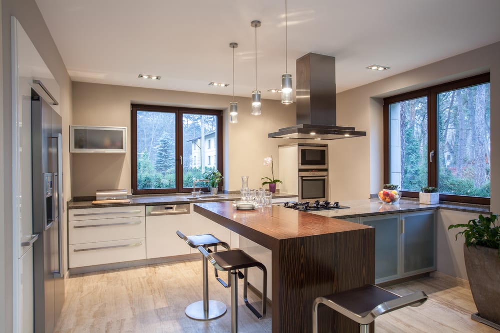 Modern kitchen with beige walls, large windows, and sleek cabinetry. A central island with dark wood countertop and bar stools under pendant lights. Cozy and inviting.