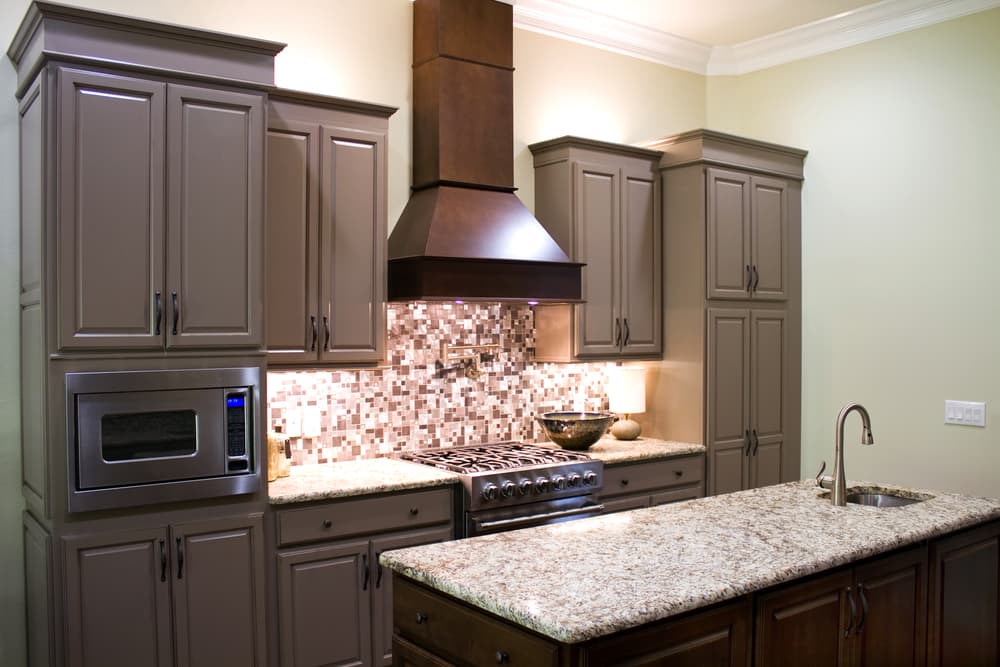 Modern kitchen with taupe cabinets, granite countertops, and stainless steel appliances. Mosaic tile backsplash and sleek vent hood above the stove.