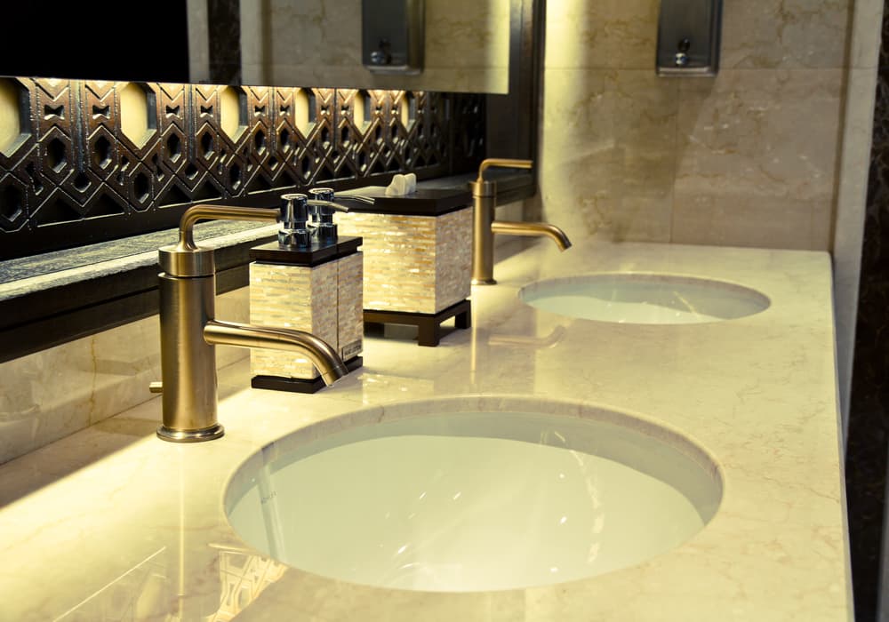 Luxurious bathroom with two round sinks in a marble counter, bronze faucets, ornate mirror, and elegant soap dispensers; warm, sophisticated ambiance.