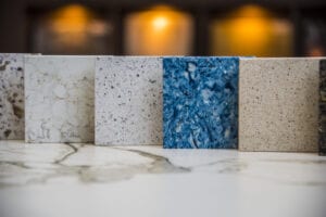 Close-up of five countertop samples in varying textures and colors—granite, marble, terrazzo, blue swirls, and beige. A blurred, warm-lit background conveys a cozy showroom ambiance.