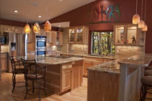 Warm-toned kitchen with wood cabinets, granite countertops, pendant lights, and bar stools. A colorful sculpture decorates the maroon wall above.