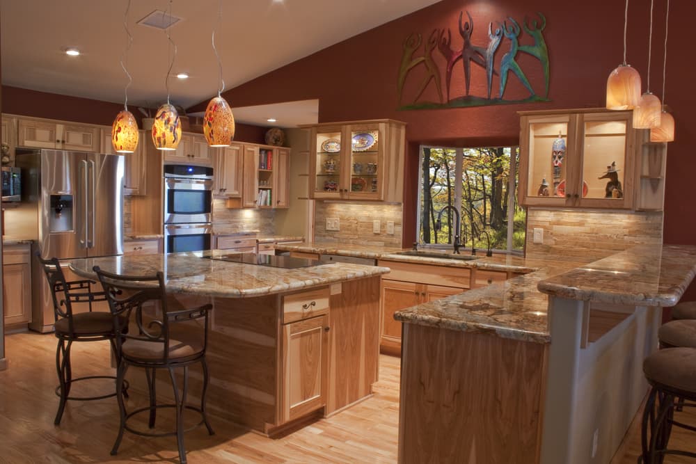 Warm-toned kitchen with wood cabinets, granite countertops, pendant lights, and bar stools. A colorful sculpture decorates the maroon wall above.