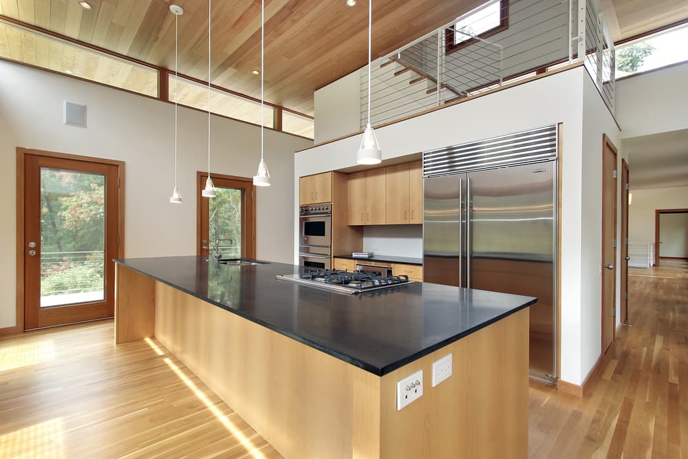 Modern kitchen with high ceilings, natural wood floors, large island with black countertop, hanging lights, stainless steel appliances, and glass door views.