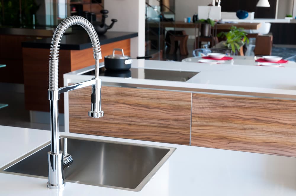 Modern kitchen with a sleek chrome spring faucet over a stainless steel sink, set within a white countertop. Wooden accents and stylish decor create a contemporary vibe.