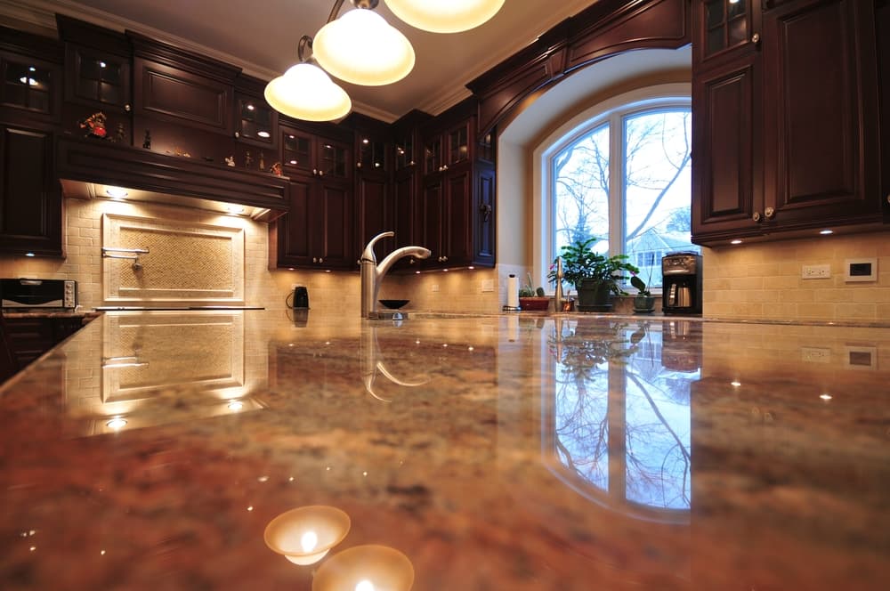 Luxurious kitchen with dark wood cabinets, granite countertop reflecting pendant lights, and a large arched window with a serene outdoor view.