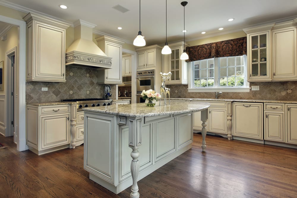 A spacious kitchen with elegant cream cabinets and a large granite island. Pendant lights hang above, while natural light pours in from a window.
