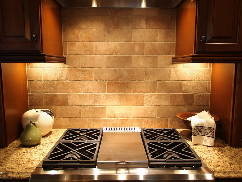 A warm, well-lit kitchen features a stone tile backsplash, black stove grates, and wooden cabinets. Decor includes vases and a neatly folded towel.