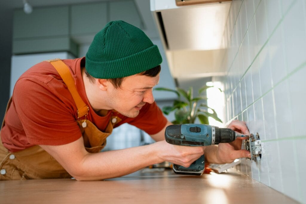 A handyman in a green beanie and brown overalls using a cordless drill to install or repair a wall outlet on a white tiled kitchen backsplash.