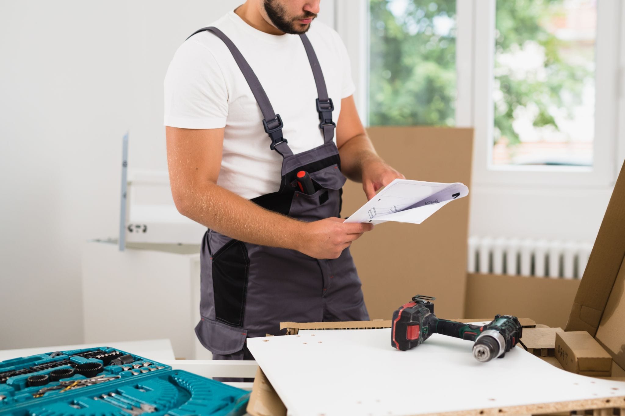 A handyman in grey overalls reviewing furniture assembly instructions in a room filled with tools and flat-pack boxes.