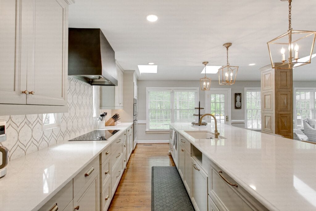 Bright, modern kitchen featuring white cabinetry, a long marble island with a gold faucet, and elegant lantern pendant lights over hardwood flooring.