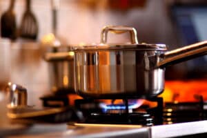 A stainless steel pot with a lid is on a lit gas stove, with a blue flame underneath. Kitchen utensils are blurred in the background, creating a warm, cozy ambiance.