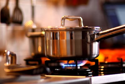 A stainless steel pot with a lid is on a lit gas stove, with a blue flame underneath. Kitchen utensils are blurred in the background, creating a warm, cozy ambiance.